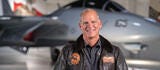 Man with a brown pilots jacket on standing in front of an airplane.
