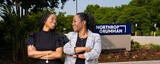 Two women in business professional dress and red heels stand, with arms crossed, in front of a Northrop Grumman facility.