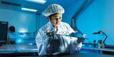 Rosibel Diaz wears a white coat, gloves and hairnet and stands at a table in a blue-lit lab, examining a mirrored panel in her hands.