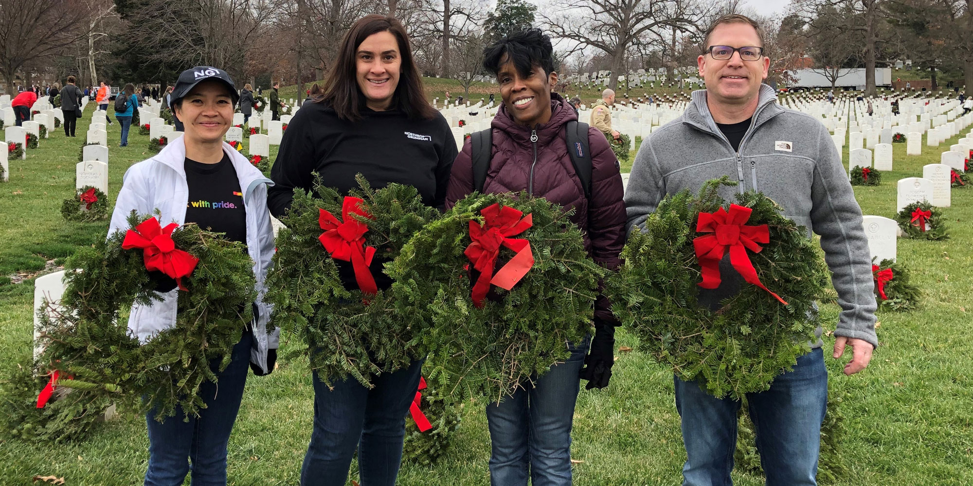 Four people standing outside in a cemetary holding holiday wreaths.