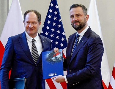 two males holding a document in front of flags