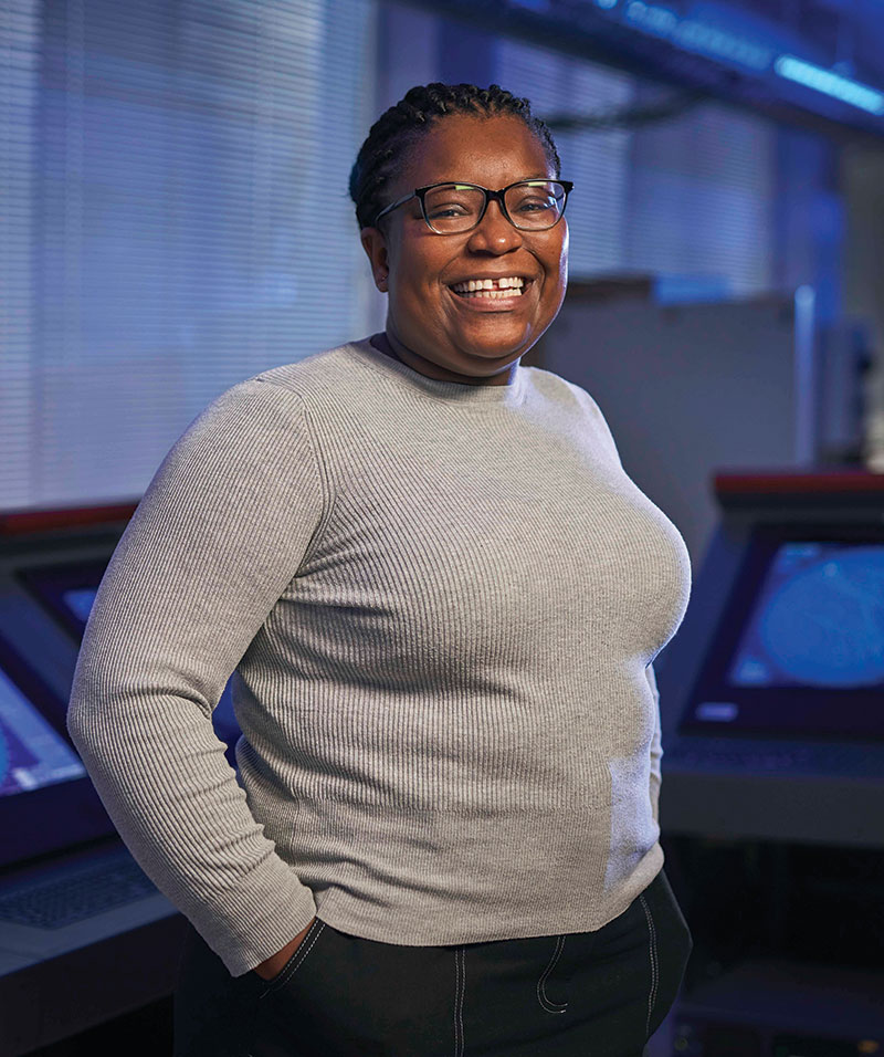 smiling female with hands in pockets standing in front of monitors