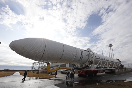 A rocket on a trailer being transported to launch pad in front of a blue, sunlit sky with light clouds