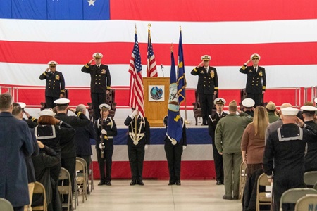 Four soldiers in dress uniforms saluting with large USA flag in background