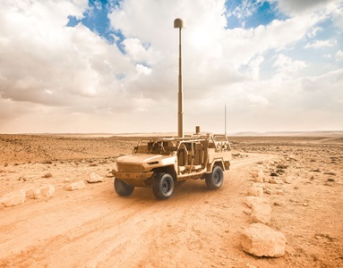 Delivering Solutions to the U.S. Army with Capacity, Scale and Speed Military desert vehicle with extended communication antenna driving on rocky dirt road under cloudy sky.