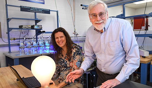 Two individuals pose smiling in front of desk