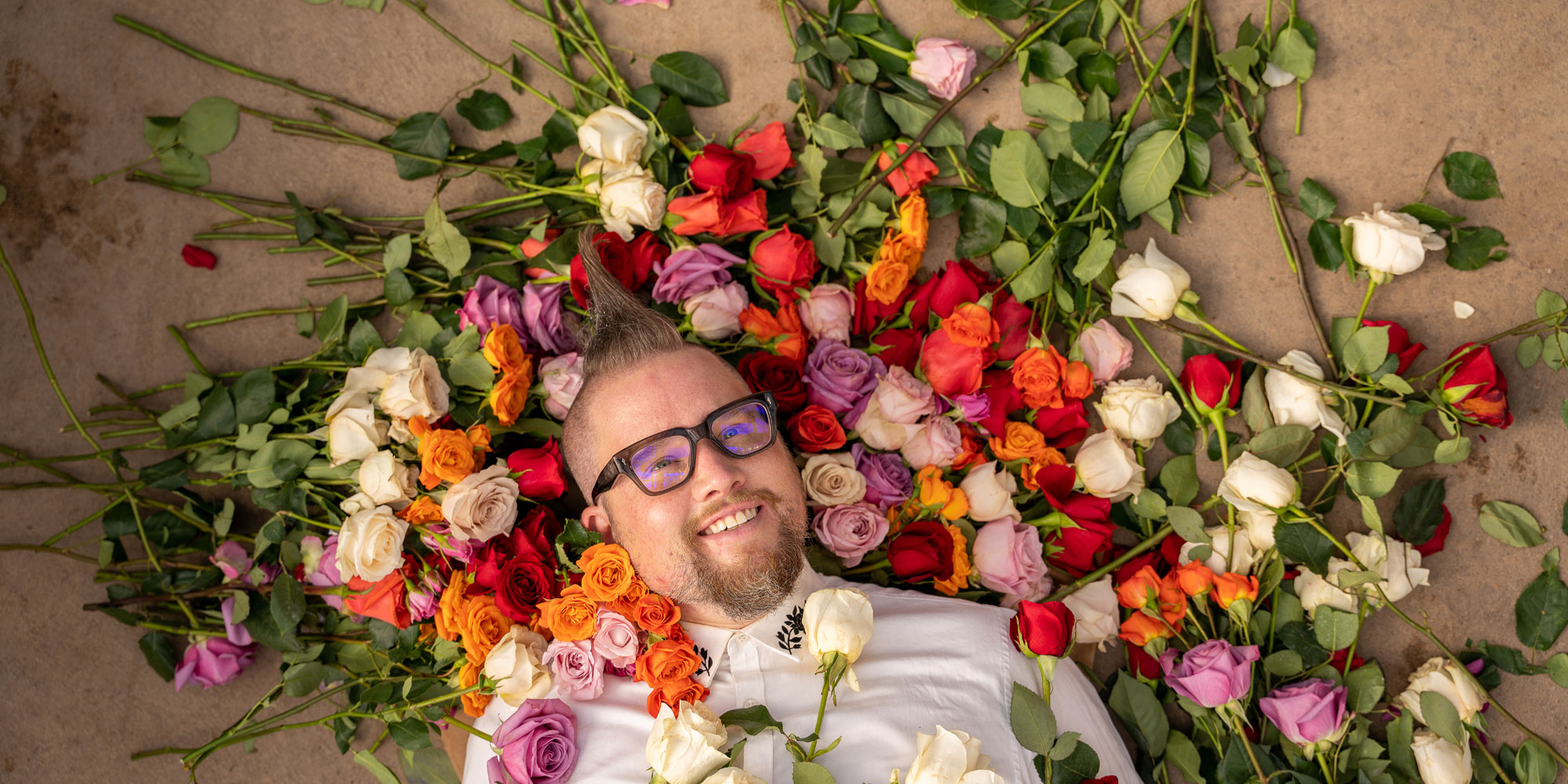 Man with mohawk and glasses wears a white button-down shirt and lays in a pile of white, pink, red and orange roses.