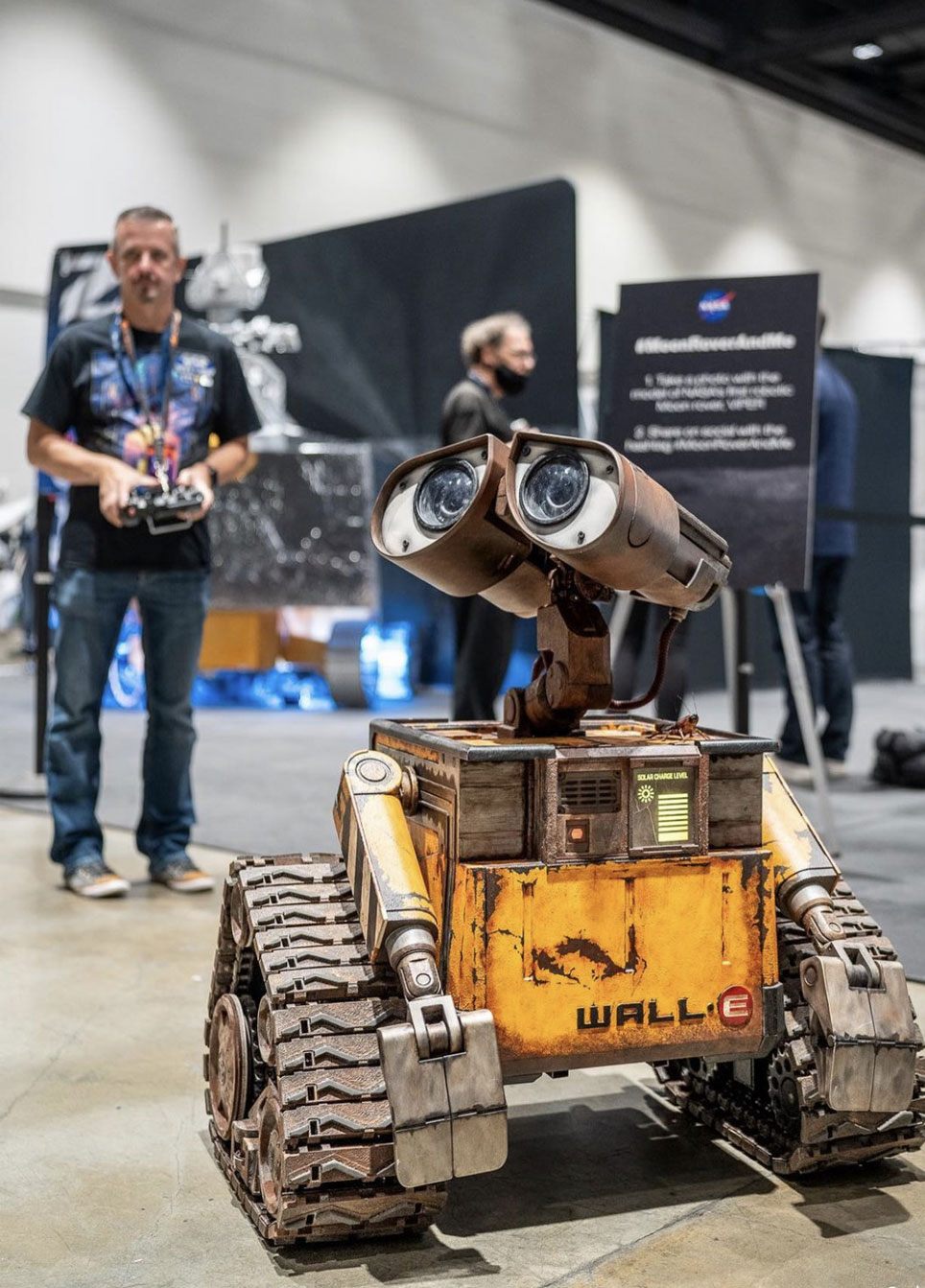 Man in grey buttondown shirt and jeans kneels next to a life-size WALL-E robot in front of the Space Shuttle