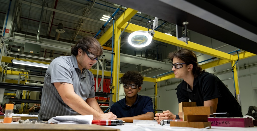 3 male employees gather around a workbench in a manufacturing area with an overhead light above them.