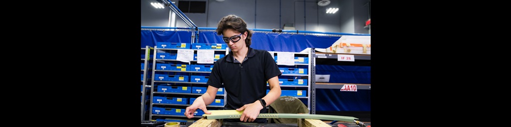 Young man with brown hair in navy polo works at a workbench.