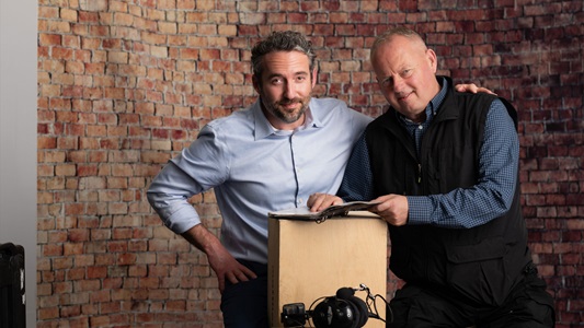 Two men stand next to each other; the man on the left wears a blue shirt and has his arm around the man on the right, who wears a button-down and sweater.