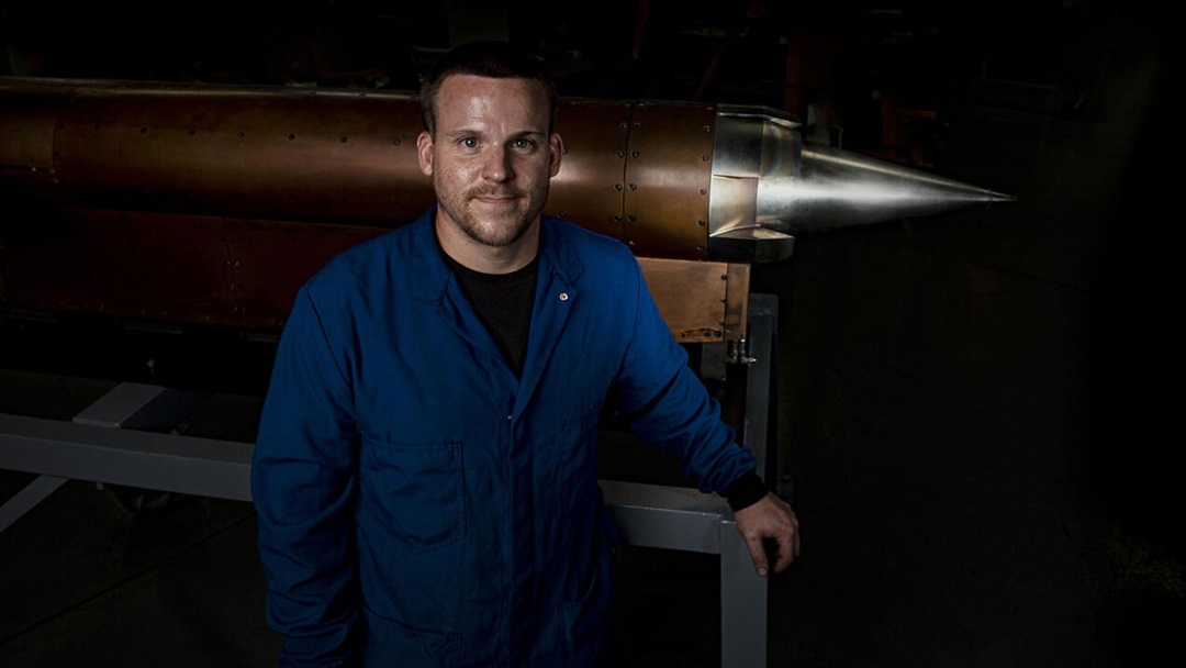 Man wearing navy jumpsuit stands in front of missile with arms crossed.