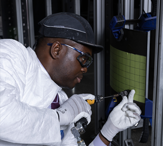 man working on equipment with white gloves, goggles, and white coat