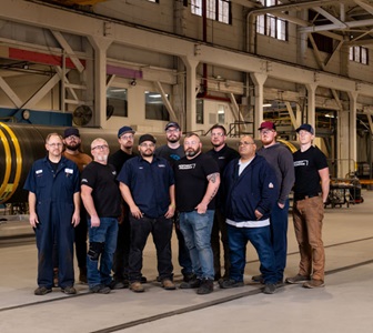 Group photo of Northrop Grumman hydrotesting team standing in a manufacturing facility, with a large solid rocket motor case in the background.  