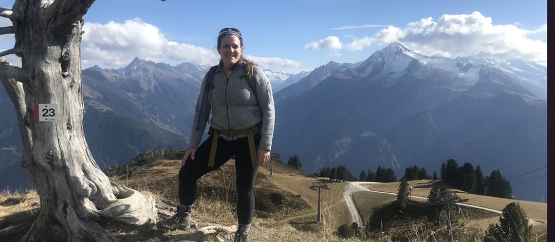 Woman dressed in outdoor gear on a hiking trail surrounded by mountain scenery. 