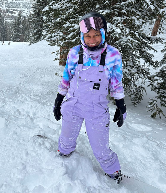 Person in purple overalls, helmet and goggles standing on a mountain top covered in snow. 