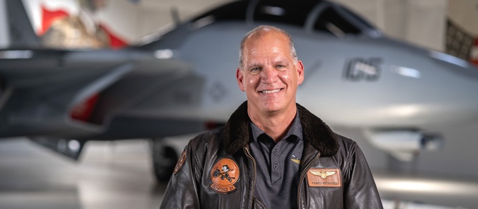 Man with a brown pilots jacket on standing in front of an airplane.  