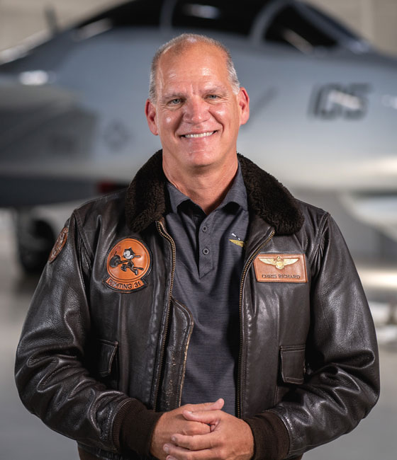 Man with a brown pilots jacket on standing in front of an airplane.  