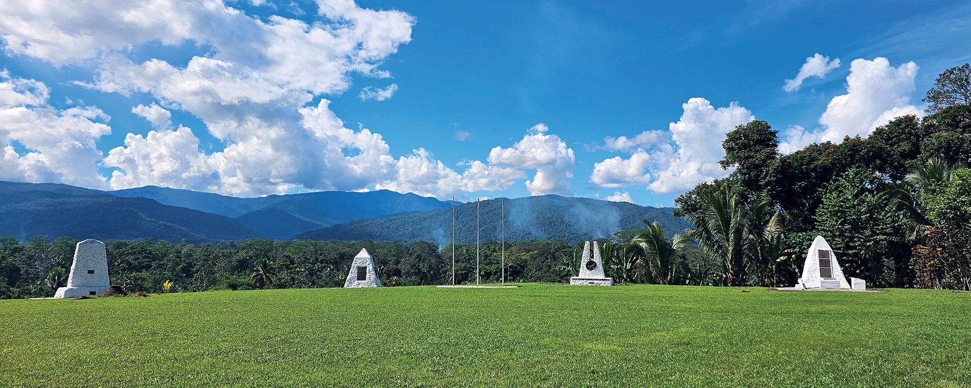 Green grass field with three white stone memorials against a jungle backdrop 