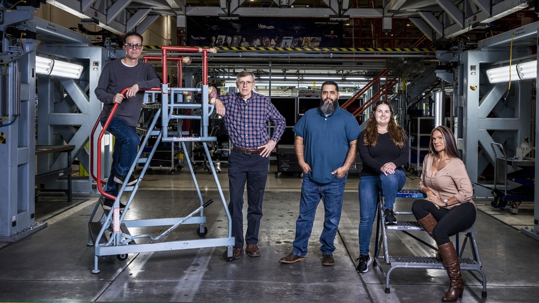 Five employees stand in an industrial space, some sitting or standing on ladders.