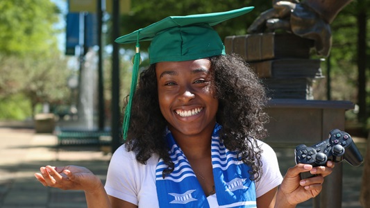 Woman in graduation cap holding game controller smiling. 