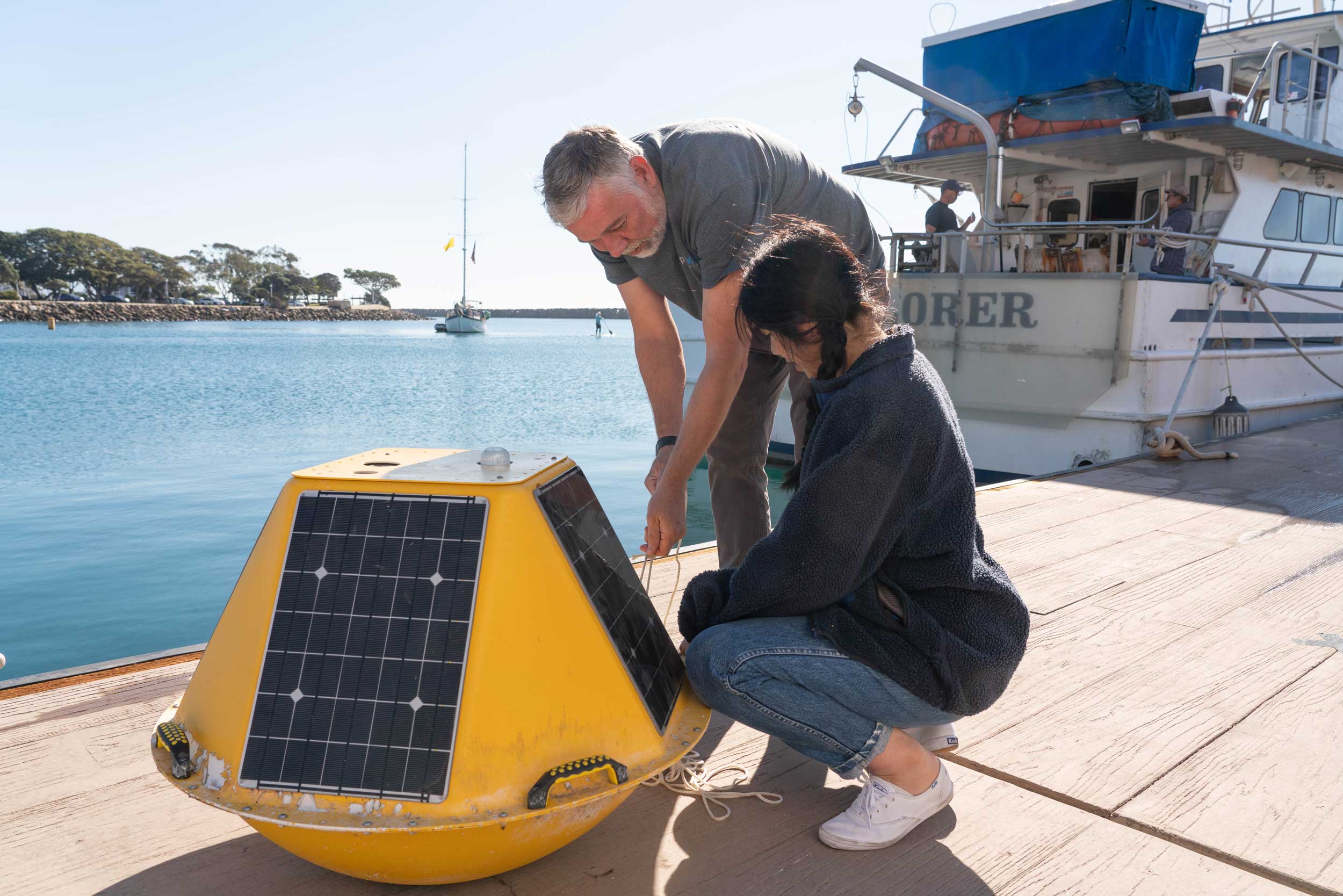 A woman and two men crouch on a dock with a yellow plastic round buoy and slim metal cylander in front of them that is the Glacier Watch system. Behind them is a red building and a boat.