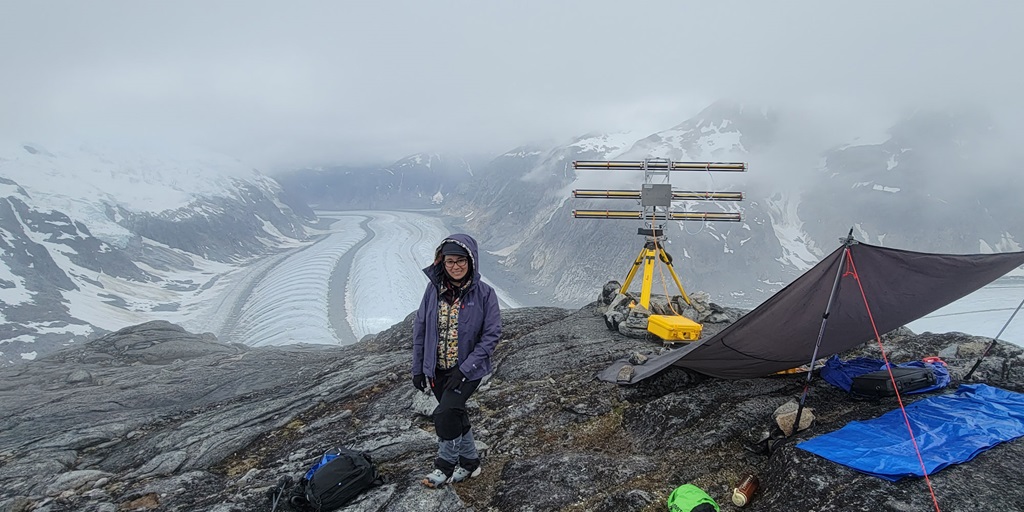 Employee Krystal wears a coat with hood over her head while standing amidst the snowy Alaska terrain with snow-covered mountains in the background.