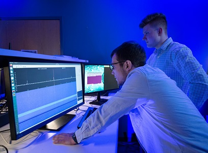 two men are standing, looking at computer screens