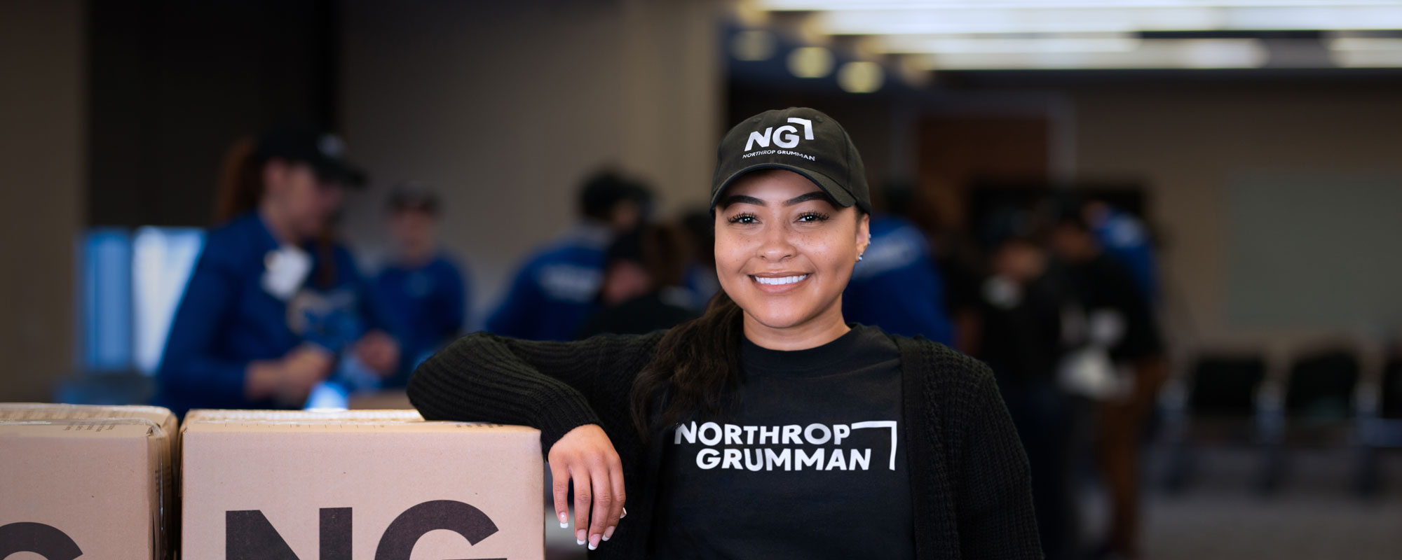 Woman smiling in Northrop Grumman shirt and cap with  items for donation. 