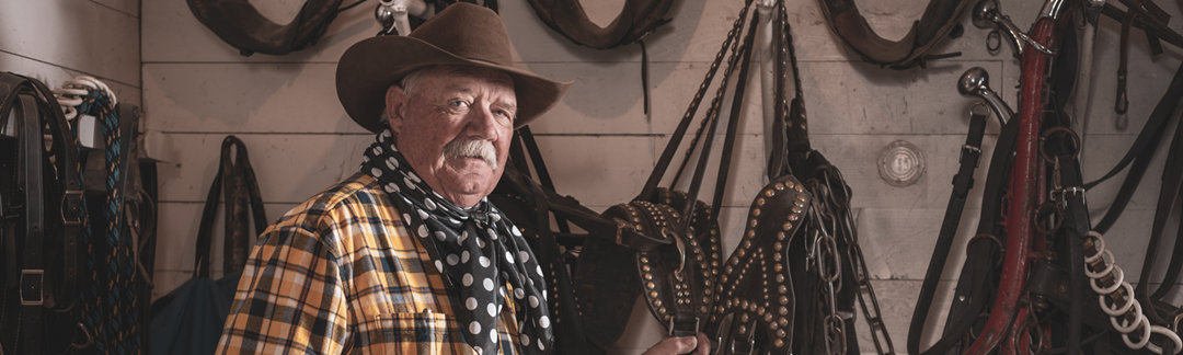 Older Caucasian man in cowboy attire poses in front of horse saddles hanging on wall