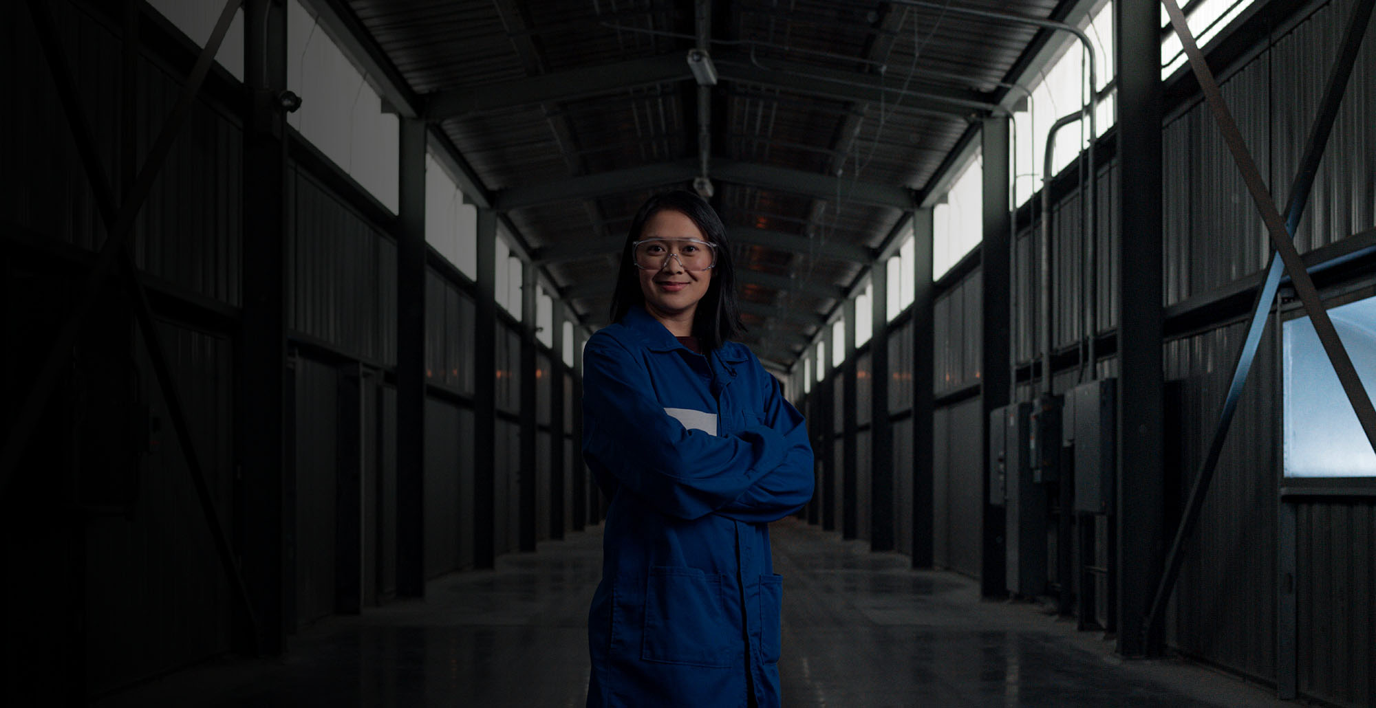 a woman wearing a blue lab coat and safety glasses with her arms crossed stands in a hallway and smiles at the camera