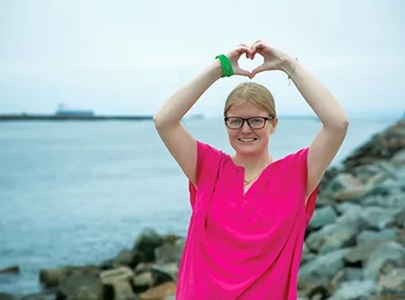 woman with glasses forms a heart wtih her hands above her head outdoors