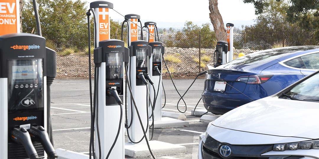 A blue and a white car parked in a lot and plugged into electric vehicle charging stations