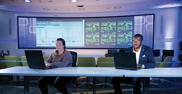 a man and woman work on laptops at a table with large computer monitors behind them