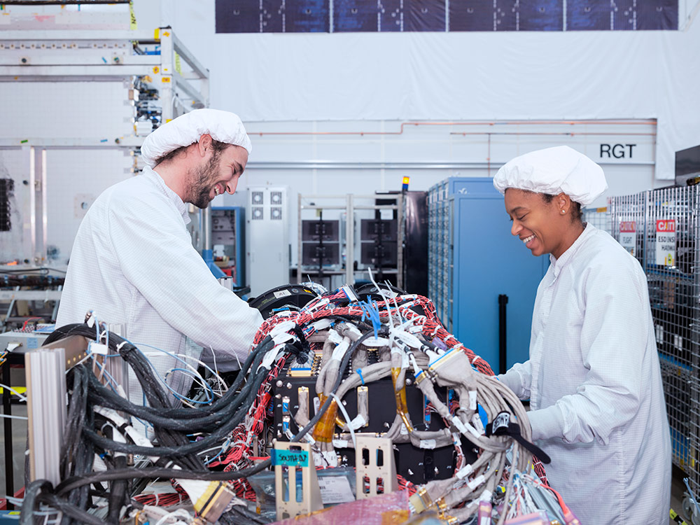 Two employees smile while working on a large machine with long wires in a lab