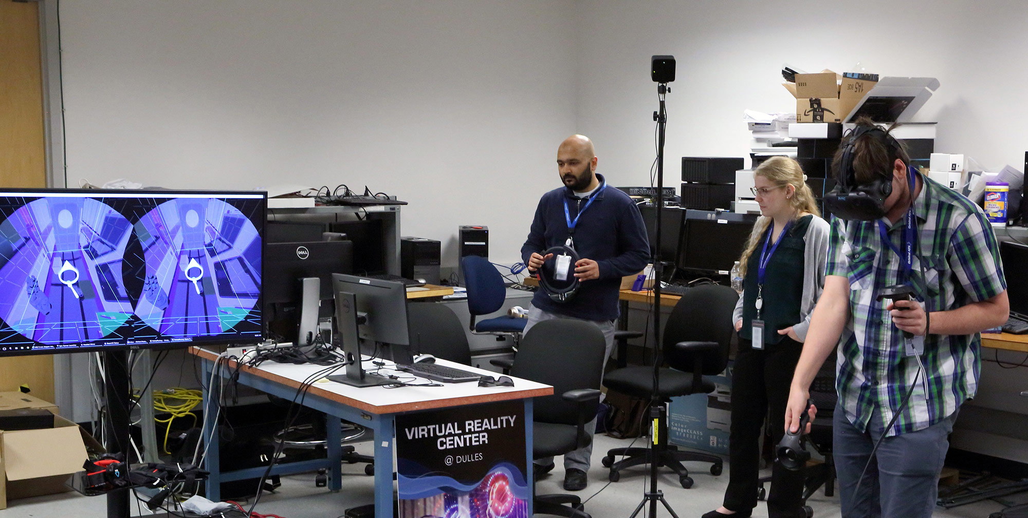 A man in a plaid shirt uses a VR headset while two engineers watch monitors in the background.