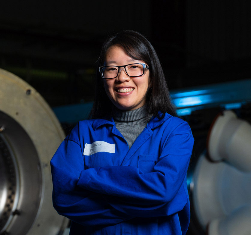 Asian female in blue lab coat and safety goggles stands in hallway of manufacturing plant