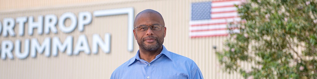 Black man standing in front of flag and Northrop Grumman logo