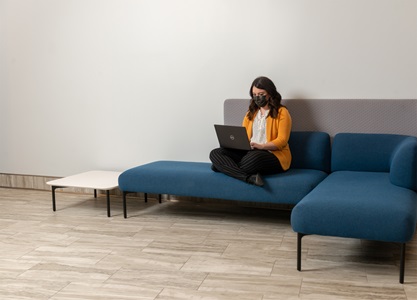 A white woman wearing a mask and dressed in a white blouse with a yellow jacket is sitting on a blue chair with a computer in her lap