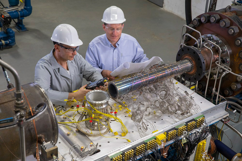 Northrop Grumman Engineers perform an instrumentation check at the hypersonics testing facility. Two men in hard hats look at instruments on a table in a manufacturing setting.