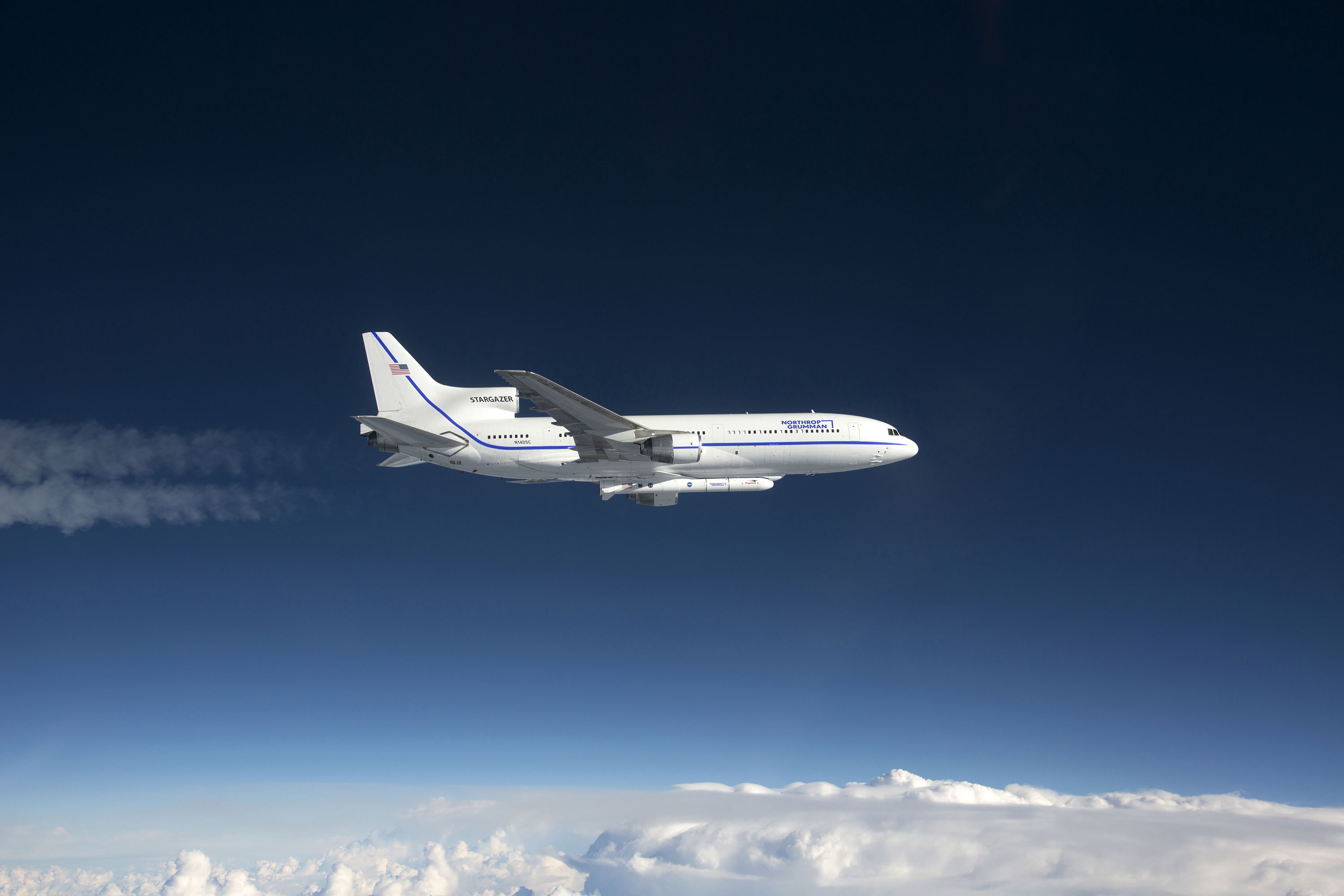 A white airplane above clouds in front of a blue sky
