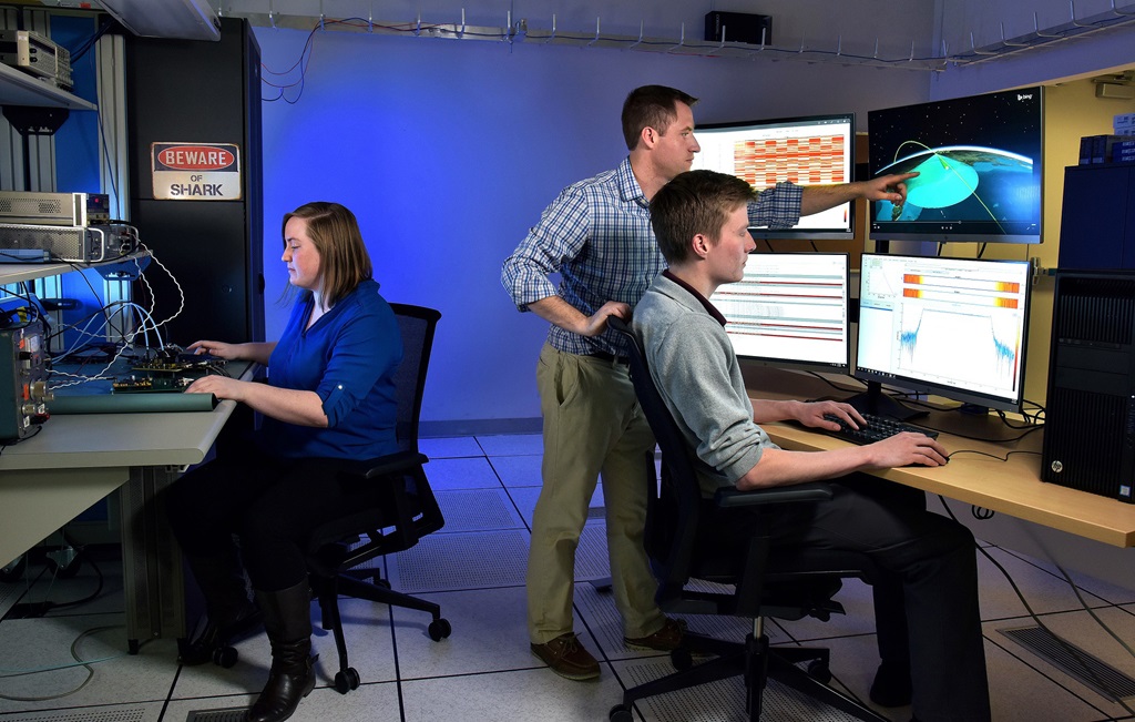 Two white males and a female in a blue lit room loooking at computer screens