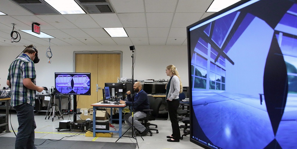 A man in a plaid shirt uses a VR headset while two engineers watch monitors in the background.