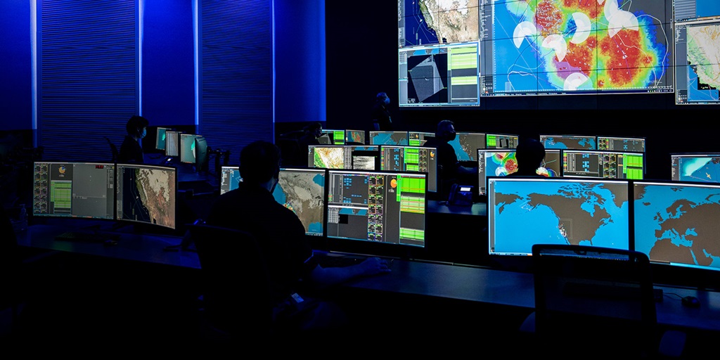 Large command center with multiple computer screens and people monitoring data.