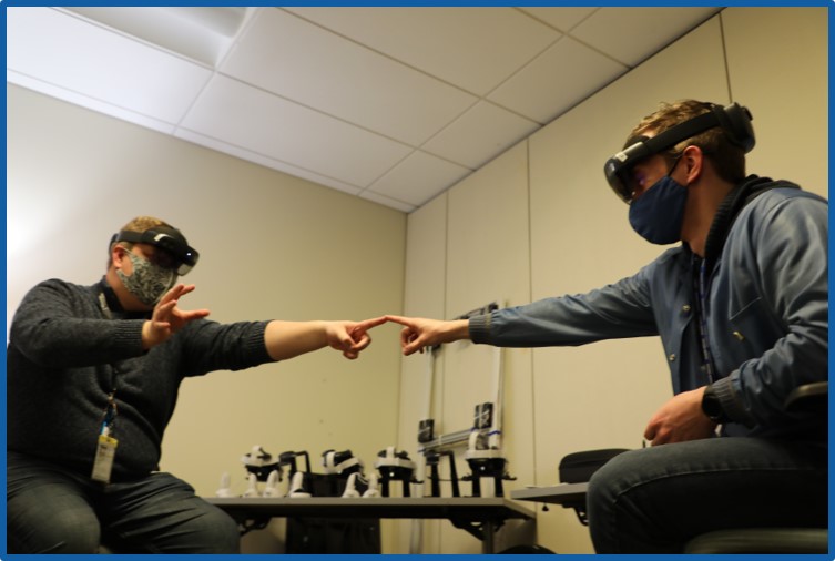 Two men wearing virtual reality headsets sit in chairs and point at each other