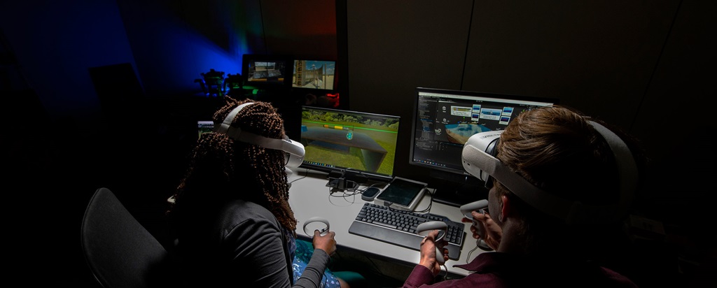 Two people sit at computers wearing virtual reality headsets