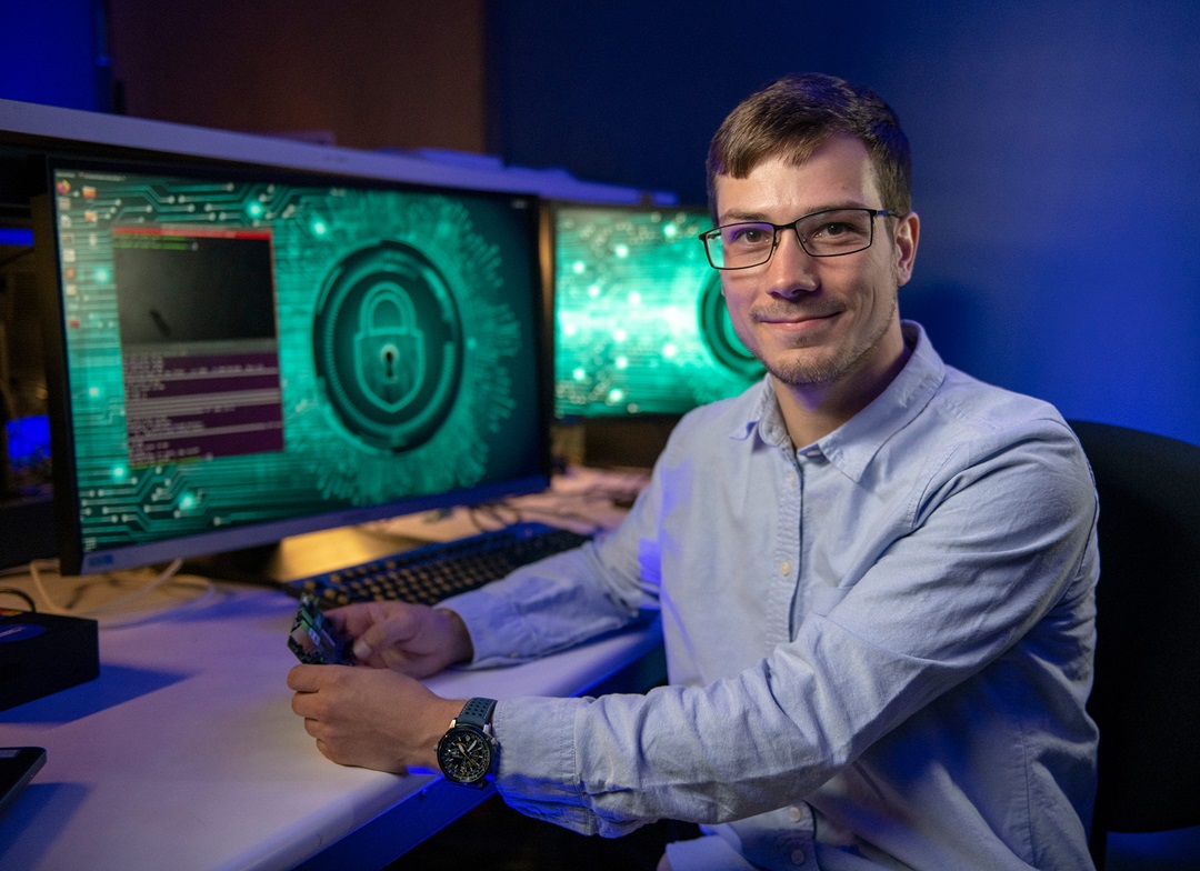 a man wearing glasses sits at dual monitors and looks directly at the camera