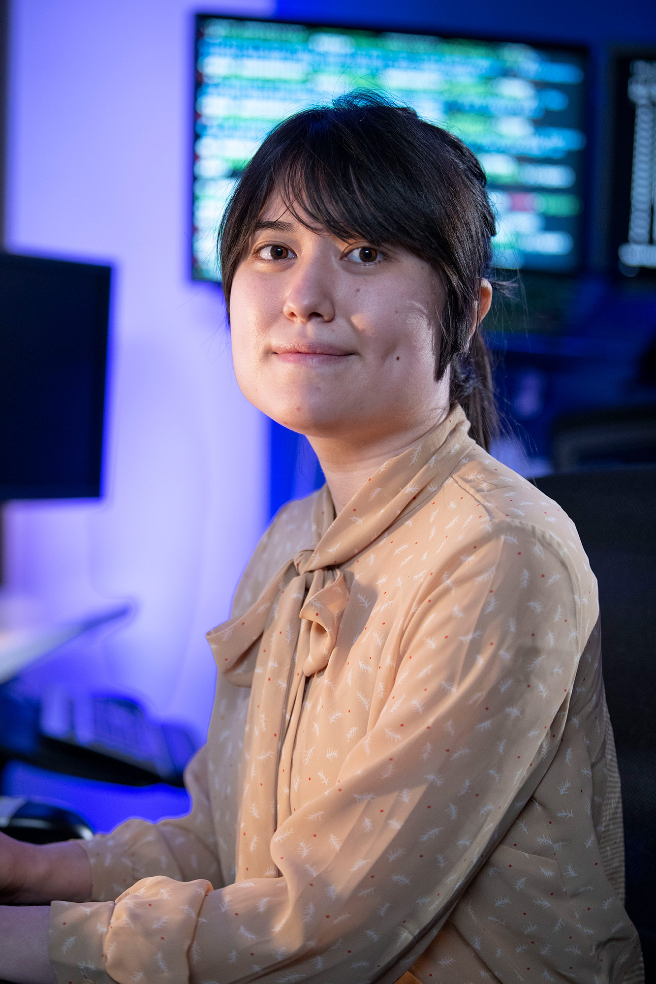 a woman looks directly at the camera with computer monitors in the background