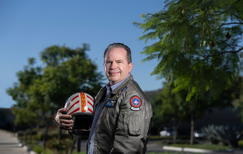 A white man wearing a "bomber" jacket and holding a helmet smiles at the camera