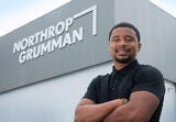 a Black male standing in a black shirt in standing in front of an office building with a Northrop Grumman sign in white letters